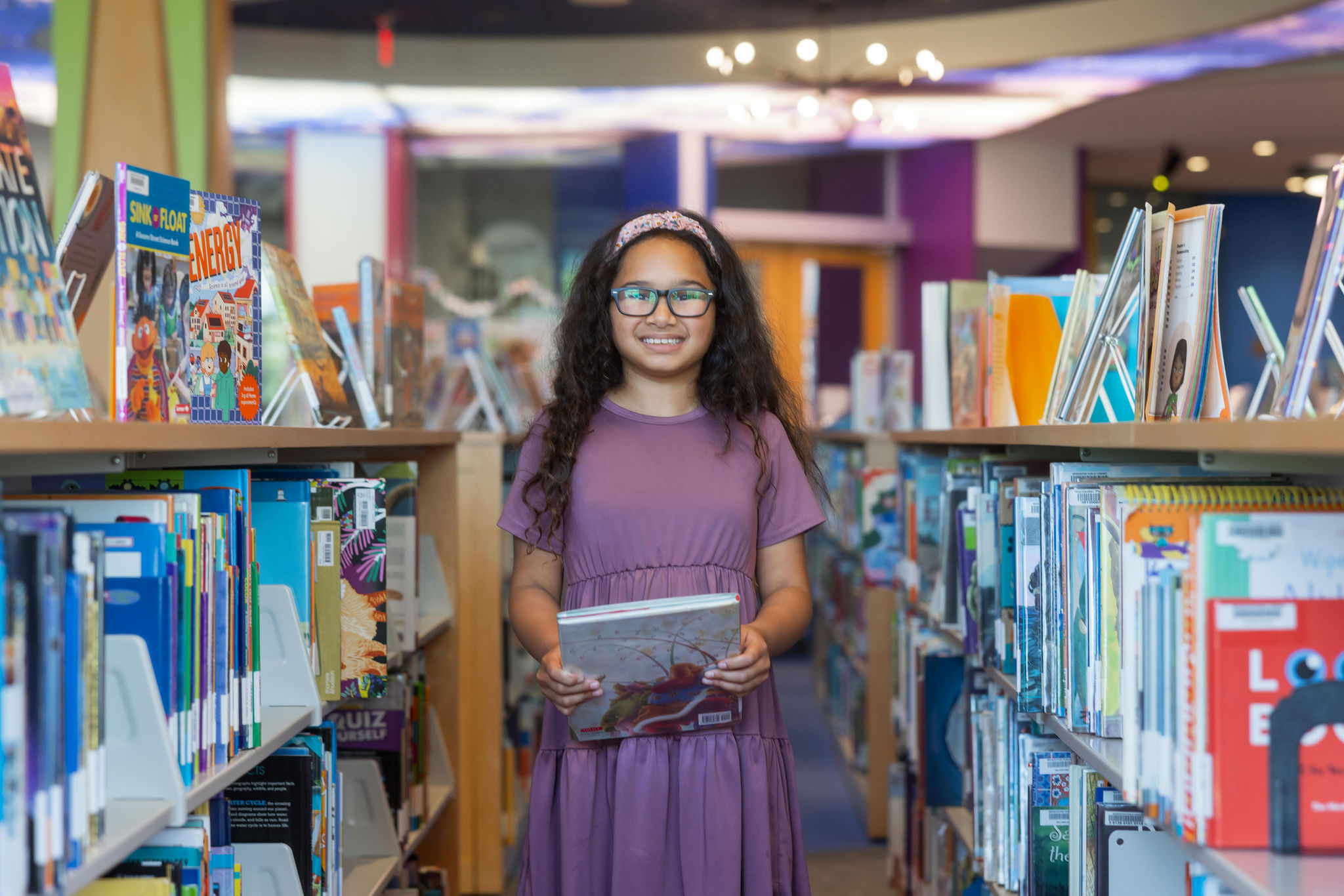 A young girl stands amongst stacks of books in the children's room while holding a book.